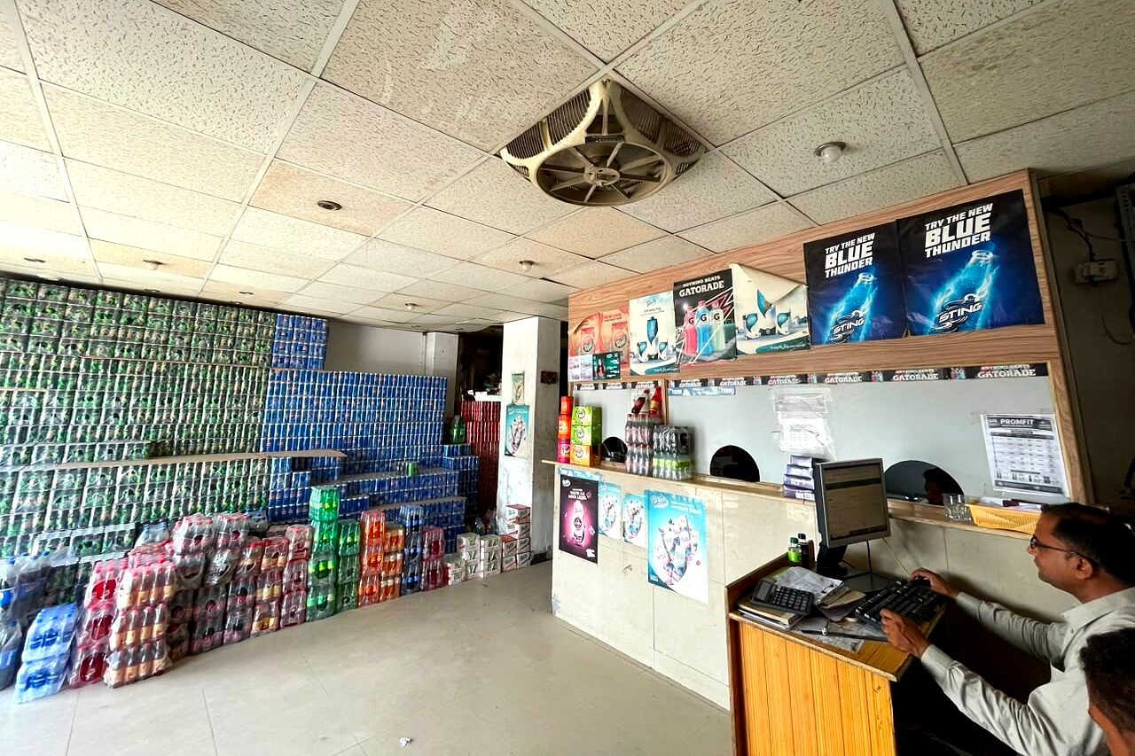 The trade counter at Buleedi Distributors, a member of staff working at the till beside the service windows, with the drinks wall stacked case on case behind.