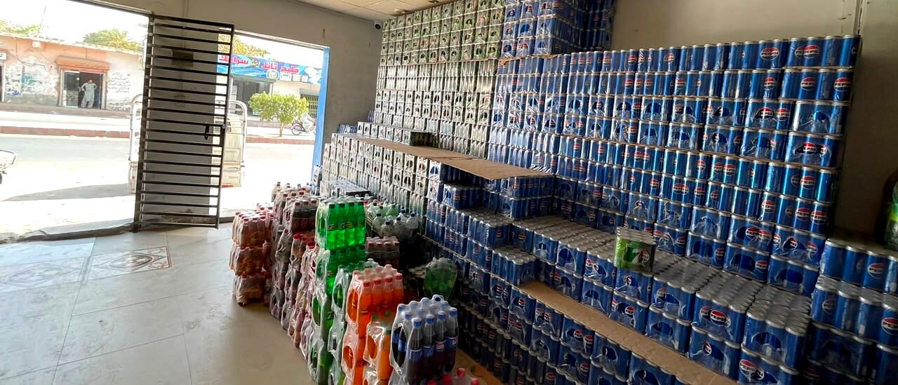 The front room at Buleedi Distributors: Pepsi cases stacked in ruled tiers from floor to ceiling along one wall, bottled lines in crates beside them, and the shutter open onto the street.