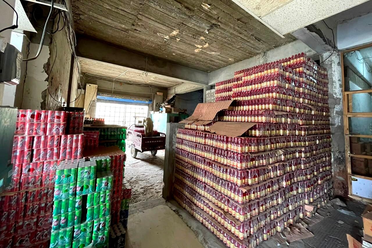 The godown at Buleedi Distributors, bottled stock shrink-wrapped and stacked in courses well above head height, with a loading rickshaw at the far doorway.